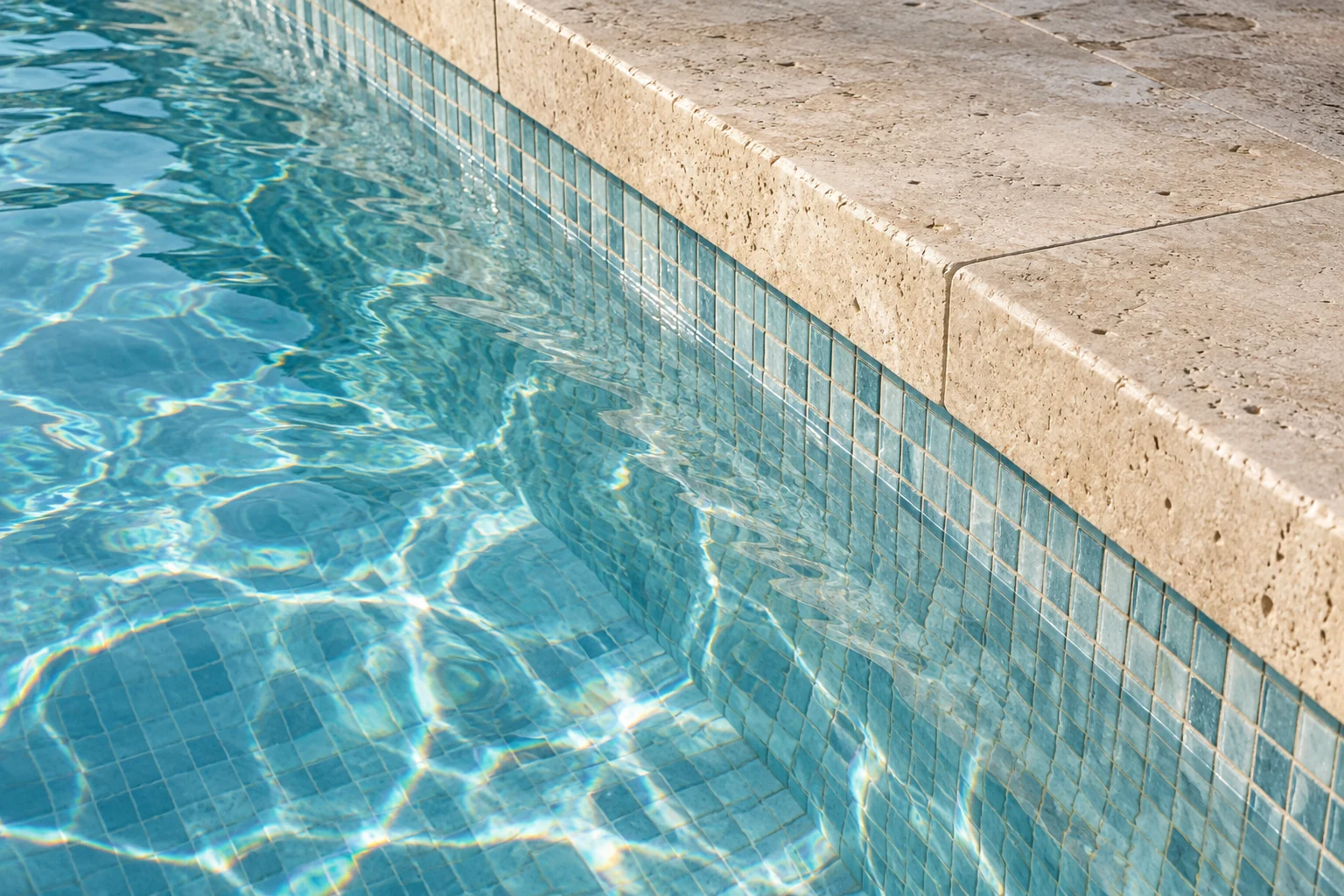 Blue mosaic beneath clear pool water beside pale stone coping.